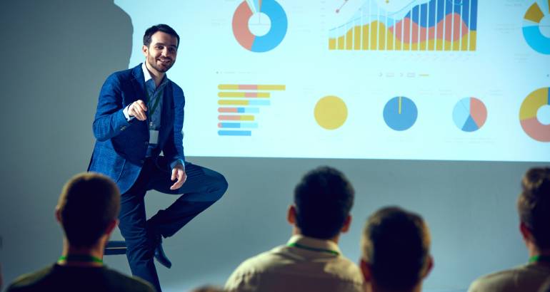 Professional trainer giving talk on data science with detailed analytical visualization projected behind him at a training room.